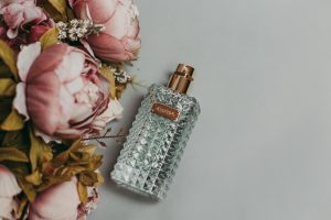 Un flacon de parfum en verre luxueux à côté de pivoines roses délicates sur un fond gris, mettant en valeur l'élégance et le parfum.