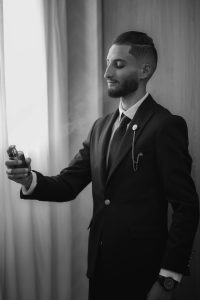 Un homme en costume élégant vaporise du parfum près d'une fenêtre ensoleillée, dégageant une impression d'élégance.