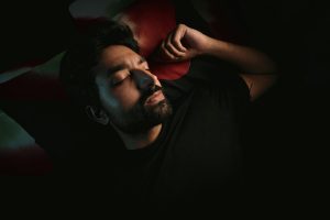 A bearded man lies in a dimly lit room, peacefully resting with closed eyes. Un homme barbu allongé dans une pièce faiblement éclairée, se reposant paisiblement les yeux fermés.